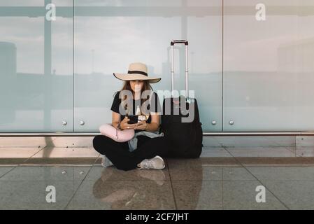 Giovane donna in cappello e abbigliamento casual seduto in lotus posa sul pavimento messaggi sullo smartphone in attesa di volo in aeroporto Foto Stock