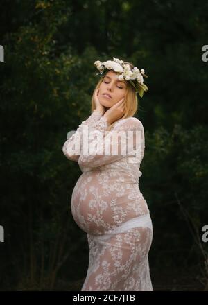 Calmo attraente donna incinta con occhi chiusi in pizzo bianco abito e bianco fiore corona sognare mentre in piedi e toccare testa tra le foglie di alberi verdi densi nella foresta in estate Foto Stock