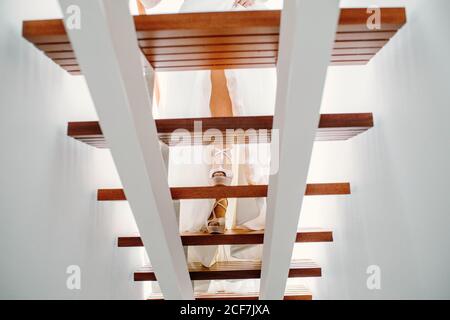 Da sotto di bride raccolto che indossa tacchi alti ed elegante abito da sposa a piedi su scala in legno in appartamento moderno Foto Stock