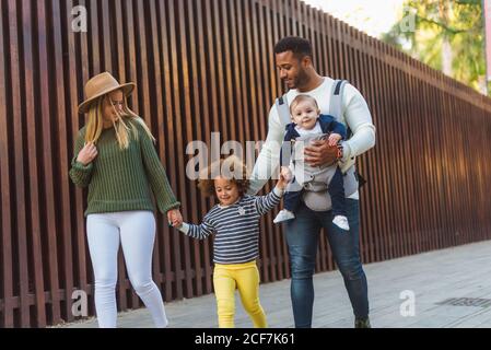 Allegro giovane coppia multirazziale di stile con bambino e poco figlia in abbigliamento casual camminando insieme lungo recinzione di legno sopra via della città