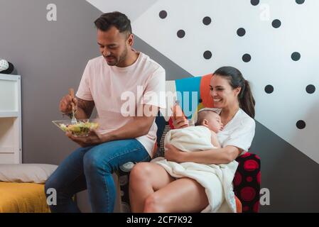 Madre dai capelli neri con petto di bebè sonnolento con mobile telefono in mano seduta su sedia con il padre in casual indossa l'insalata da mangiare Foto Stock