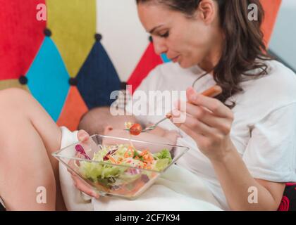 Vista laterale della giovane madre dai capelli neri in un abbigliamento casual seduto su poltrona e allattando il bambino mentre si mangia insalata sana Foto Stock