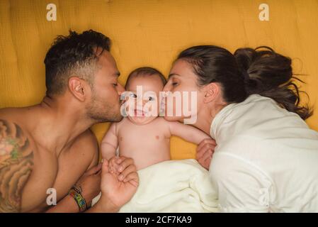 Vista dall'alto di giovani genitori felici che baciano in guance poco bambino soddisfatto mentre si sdraiano sul letto Foto Stock