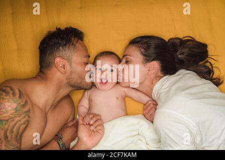 Vista dall'alto di giovani genitori felici che baciano in guance poco bambino soddisfatto mentre si sdraiano sul letto Foto Stock