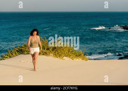 Tanned slim Donna in pantaloncini bianchi a piedi nudi sulla spiaggia sabbiosa con pianta sullo sfondo di onde turchesi schiumose a Fuerteventura, Las Palmas, Spagna Foto Stock