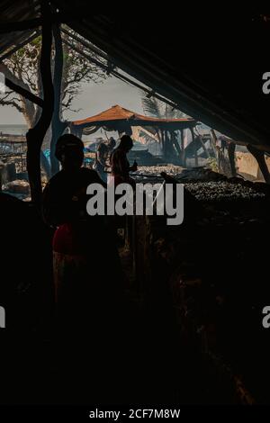 Gambia, Africa - 3 agosto 2019: Donna africana irriconoscibile in ombra che smista il pesce dal paniere vicino al bancone sotto la tenda oscura del mercato di strada Foto Stock