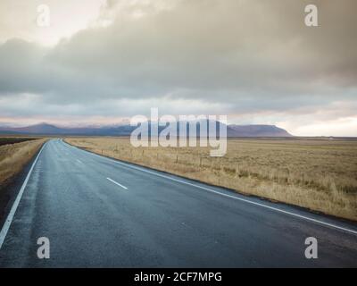 Pittoresca vista di percorso asfaltato che conduce tra enormi prati con Erba secca vicino alle colline e cielo nuvoloso in Islanda Foto Stock