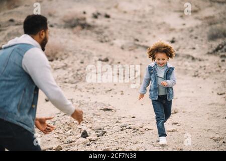 Allegro uomo etnico bearded vestito con abiti eleganti giocando con bimbo etnico riccamente felice sulle colline di sabbia chiara di giorno Foto Stock