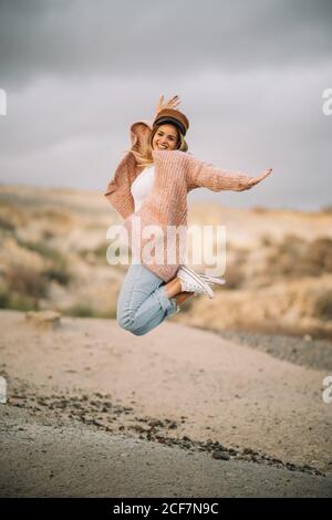 Femmina sorridente con capelli biondi vestiti eleganti e. cappello che salta con le braccia allungate e le gambe piegate terra sabbiosa e guardando la macchina fotografica Foto Stock