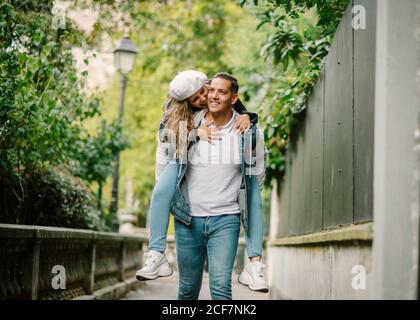 Content Donna in abbigliamento casual seduta sulla schiena e baciando l'uomo compiaciuto a piedi su un piccolo vicolo con piante verdi su sfondo sfocato Foto Stock