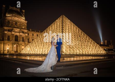 Giovane coppia appena sposata in abito da sposa e abito hugging Mentre si sta in piedi in arco a dondolo con Louvre sullo sfondo a. Parigi Foto Stock