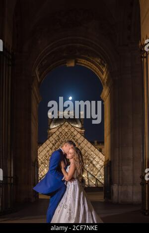 Giovane coppia appena sposata in abito da sposa e abito hugging Mentre si sta in piedi in arco a dondolo con Louvre sullo sfondo a. Parigi Foto Stock