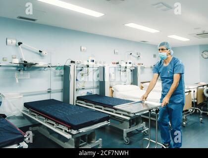 Medic in uniforme blu e vassoio di impostazione della maschera protettiva attivato trolley in camera d'ospedale da letti vuoti Foto Stock