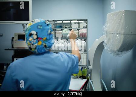 Vista posteriore della medica femminile in uniforme blu che prende i rifornimenti necessario per la chirurgia in ospedale Foto Stock