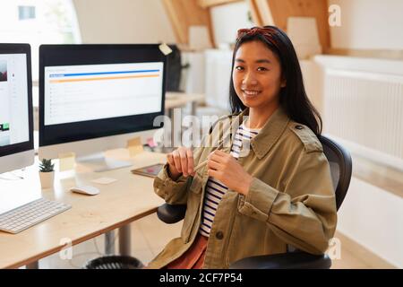 Ritratto di giovane designer asiatico seduto sulla sedia di fronte del suo posto di lavoro con i computer e sorridendo alla macchina fotografica Foto Stock