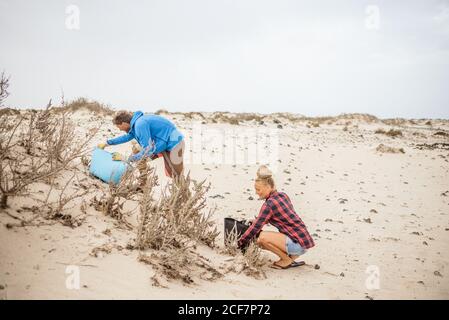 Hippster positivo Donna e uomo in abiti casual e guanti raccogliendo rifiuti in borsa mentre squatting sulla spiaggia deserta Foto Stock