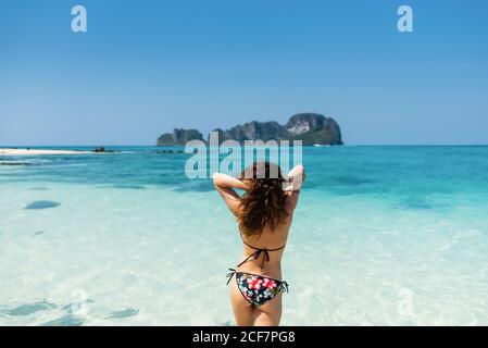 Vista sul retro della slim femminile turistica in costume da bagno in piedi Costa sabbiosa di oceano turchese sotto il cielo blu in Thailandia Foto Stock