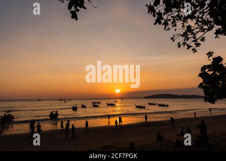 Silhouette di persone irriconoscibili che camminano sulla spiaggia di sabbia e barche Sul mare ondulato durante il bellissimo tramonto in Thailandia Foto Stock