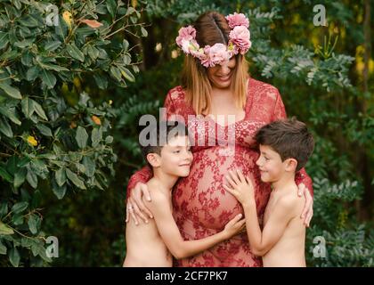 Madre felice in bel vestito rosso e corona floreale sorridente e abbracciando i due figli gemellati che coccolano il ventre mentre si sta in piedi vicino al verde alberi in giardino durante il giorno d'estate Foto Stock