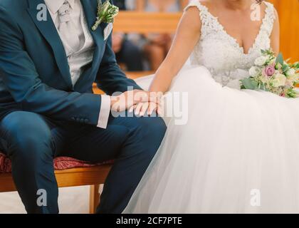 sposo irriconoscibile in abito elegante con boutonnière e sposa in abito da sposa bianco seduto sulla panca e tenendo le mani durante cerimonia in chiesa Foto Stock