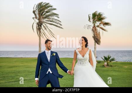 Sposo allegro in abito elegante e sposa in matrimonio bianco abiti coccolarsi e camminare sul prato guardando ciascuno altro durante la celebrazione del matrimonio Foto Stock