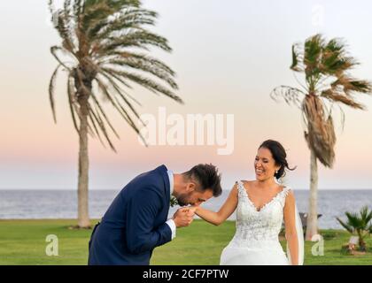 Sposo allegro in abito elegante e sposa in matrimonio bianco abiti coccolarsi e camminare sul prato guardando ciascuno altro durante la celebrazione del matrimonio Foto Stock