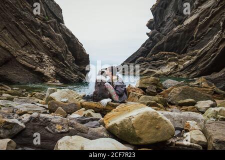 Vista laterale della donna in abiti casual seduti accanto al confine Collie e baciare cane sulla costa rocciosa contro gola e. acque tranquille della baia sotto il cielo nuvoloso Foto Stock