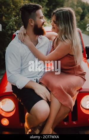 Tenero uomo barbuto in camicia bianca e pantaloncini neri che abbraccia sorridente Donna in vestito mentre si siede sul cappuccio rosso dell'automobile sull'autostrada vicino alla foresta verde Foto Stock