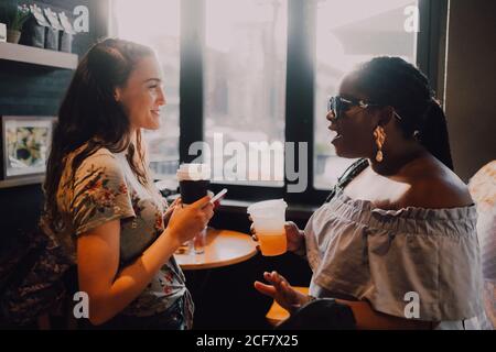 Vista laterale di allegre donne casual multirazziale e ridere bere caffè mentre si siede vicino alla finestra al caffè al tramonto Foto Stock