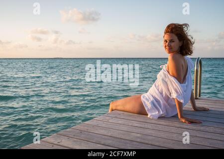 Vista laterale di una bella giovane femmina sorridente e guardando macchina fotografica mentre si siede sul molo di legno vicino all'acqua di mare increspata Contro il cielo nuvoloso sera alle Maldive Foto Stock