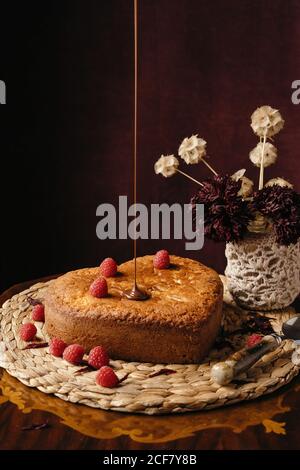 Appetitosa torta fatta in casa con bacche fresche versate con cioccolato guarnitura posta sul vassoio di vimini sul tavolo con fiori Foto Stock