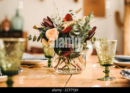 Bouquet di fiori vari e ramoscelli di piante verdi in vaso con acqua su un tavolo di legno per un pasto Foto Stock