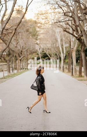 Vista laterale di un'elegante donna d'affari con gonna in pelle nera e giacca che attraversa il percorso asfaltato nel parco cittadino mentre si viaggia per lavoro Foto Stock