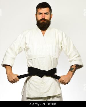 Concetto di addestramento e combattimento. Uomo con barba in kimono bianco su sfondo bianco. JUDO master tiene e legato cintura nera. Karate uomo con viso serio in uniforme. Foto Stock