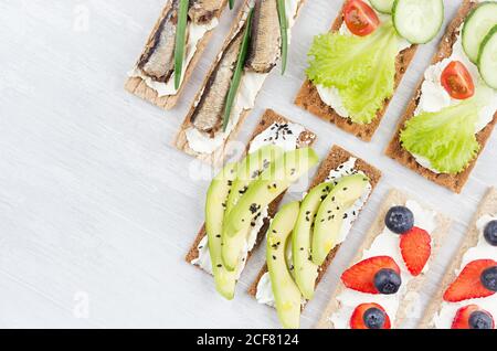 Set di panini di stagione biologica su croccanti a grani marroni secchi con verdure fresche, frutta matura, conserve di pesce, formaggio cremoso su tavolo di legno bianco Foto Stock
