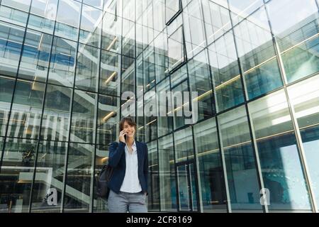 Angolo basso di felice donna d'affari che tiene la mano in tasca e usa lo smartphone mentre si trova fuori dal centro affari sulla strada della città Foto Stock