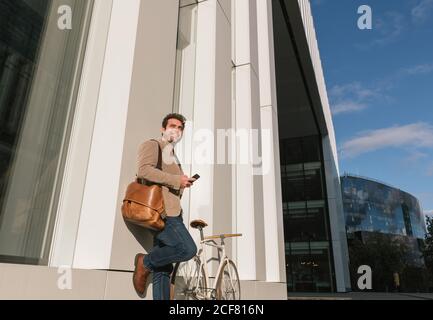 Basso angolo di imprenditore serio con smartphone appoggiato alla parete di un edificio moderno e che guarda lontano in centro Foto Stock