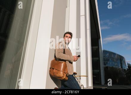 Basso angolo di imprenditore serio con smartphone appoggiato alla parete di un edificio moderno e che guarda lontano in centro Foto Stock