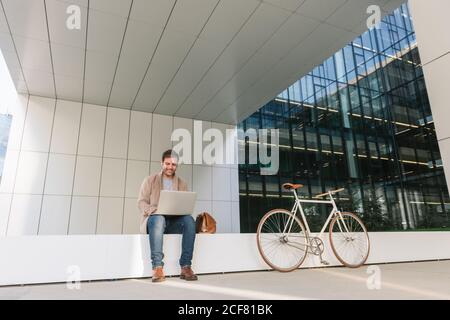 Full Body uomo felice manager sorridendo e navigando computer portatile mentre seduto all'esterno vicino alla bicicletta in un edificio contemporaneo in città via Foto Stock