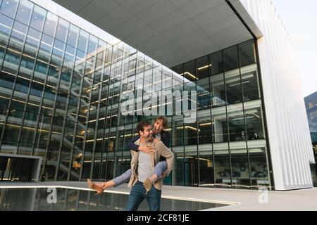 Felice uomo adulto in abiti casual trasportare Donna piggyback mentre si è in piedi e ridere sulla strada contro centro business contemporaneo in centro Foto Stock