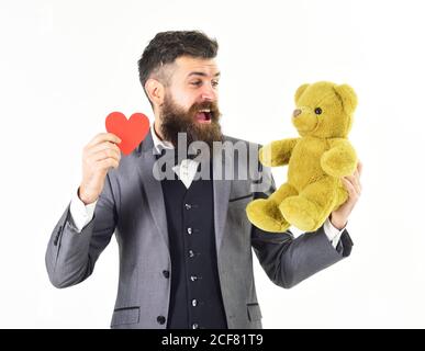 L'uomo si prepara per la data. L'uomo in costume con il volto sorridente tiene un orso peluche. Gentleman con barba e baffi tiene il cuore rosso o carta valentine, isolato su sfondo bianco. Concetto di giorno di San Valentino. Foto Stock