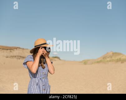 Donna adulta in cappello di paglia e vestito con macchina fotografica scattare foto sulla duna di sabbia della spiaggia in una giornata di sole Foto Stock