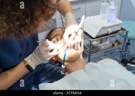 Da sopra specialista sicuro in guanti chirurgici che fanno l'iniezione con siringa e lavoro con utensili dentali in bocca del paziente sdraiato sotto la luce luminosa nella sedia nel mobile Foto Stock