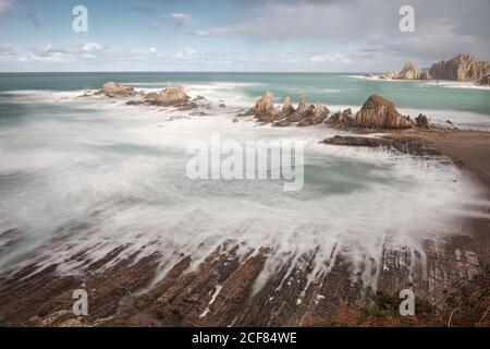 Dall'alto acqua turchese chiara e turbolente onde di schiuma bianca Sulla spiaggia di la Gueirua con incredibile erosione in composizione con forte formazioni rocciose sotto il cielo blu coperto da nubi soffici In Spagna Foto Stock