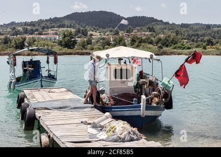 Vista laterale della donna matura che passa dal molo di legno mentre sali a bordo di un vecchio yacht colorato in acque calme, Grecia Foto Stock