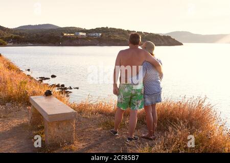 Vista posteriore di uomo e donna anziani abbronzati in cappelli godendo di splendide viste abbracciando il tramonto sul mare a Halkidiki Foto Stock