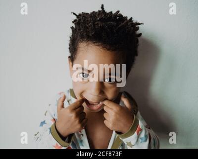 Adorabile afro-americano ragazzo in pigiama guardando la macchina fotografica e. faccia divertente mentre si è in piedi contro parete grigia Foto Stock