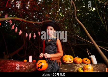 Ritratto di bambina in costume di strega seduta tra zucche E guardando la fotocamera durante la festa di Halloween Foto Stock