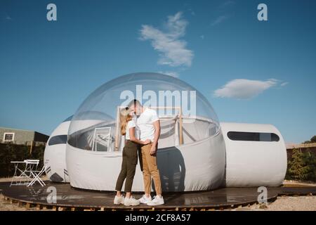 Vista laterale dell'appassionato uomo che tiene e bacia la giovane donna in piedi di fronte al romantico glamping trasparente Foto Stock