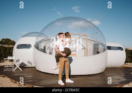 Vista laterale dell'appassionato uomo che tiene e abbraccia la giovane donna mentre si trova di fronte a un romantico glamping trasparente Foto Stock